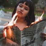 A woman stands outdoors holding a burning newspaper, capturing a dramatic and intense moment.